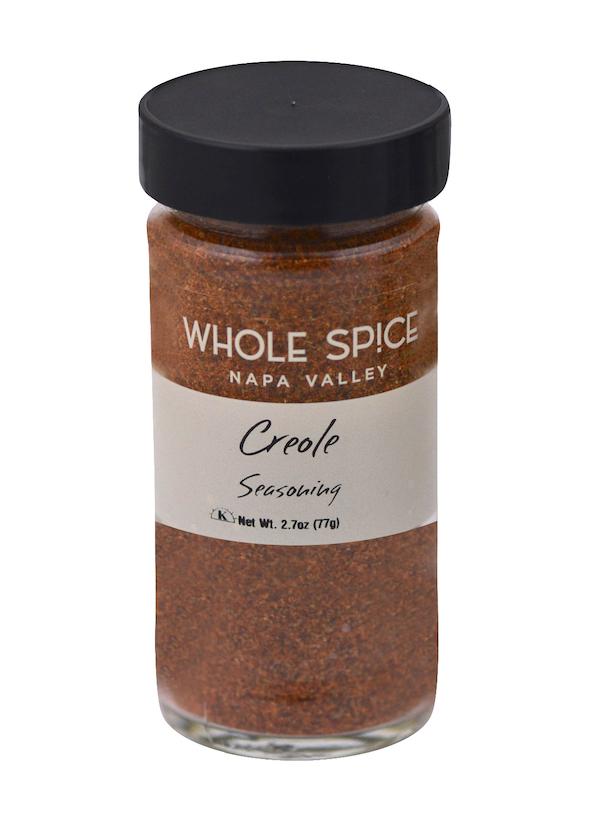 A clear glass jar with a black lid from Whole Spice Inc., labeled Creole Seasoning, holds 2.7 oz (77g) of a reddish-brown versatile blend inspired by New Orleans flavors.