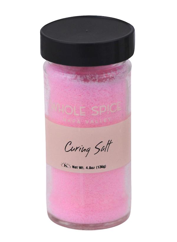 A clear glass jar with a black lid holds bright pink curing salt by Whole Spice Inc., ideal for pickling or preserving meats. The label says Curing Salt and shows a net weight of 4.8 oz (136g).