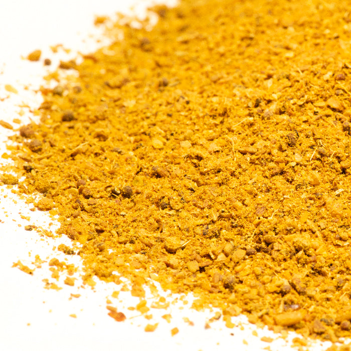 A close-up of Whole Spice Inc. Madras Curry Coarse shows its vibrant yellow powder with visible spices and seeds scattered on a white surface.