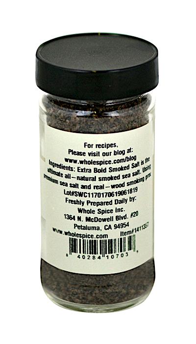 A clear jar with a black lid holds dark granules of Whole Spice Inc. Extra-Bold Smoked Sea Salt. The label lists ingredients, online recipes, and the manufacturer's address in Petaluma, California.