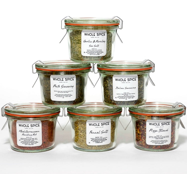 Six Weck jars from Whole Spice Inc.'s Small Mediterranean Gift Set, each labeled with a different blend—including garlic & parsley sea salt, Italian seasoning, and pizza blend—are stacked in a pyramid against a white background.