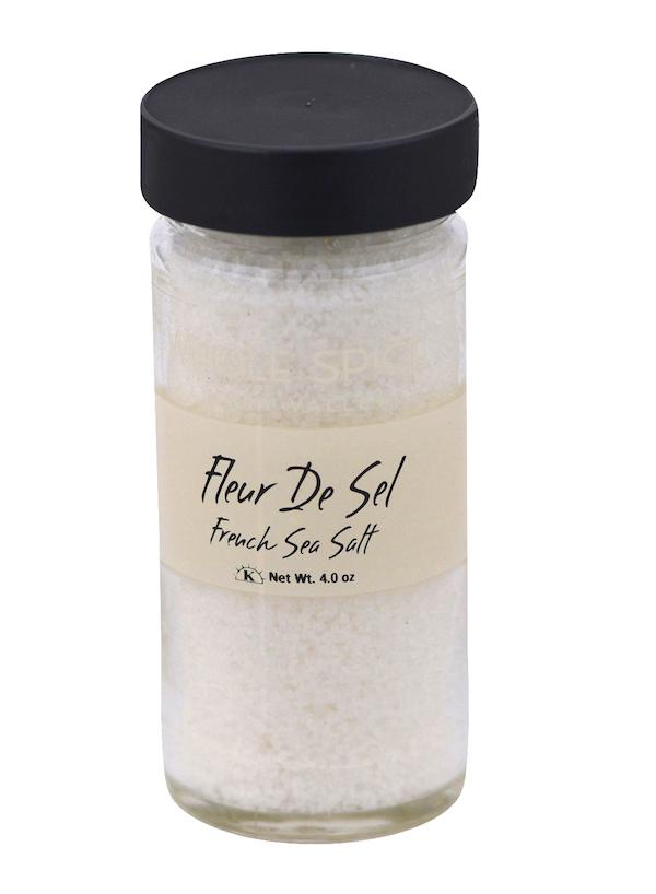 A clear glass jar with a black lid contains Whole Spice Inc. Fleur de Sel French Sea Salt. The label reads "French Sea Salt, Net Wt. 4.0 oz" in black text, and the salt inside is white finishing salt.