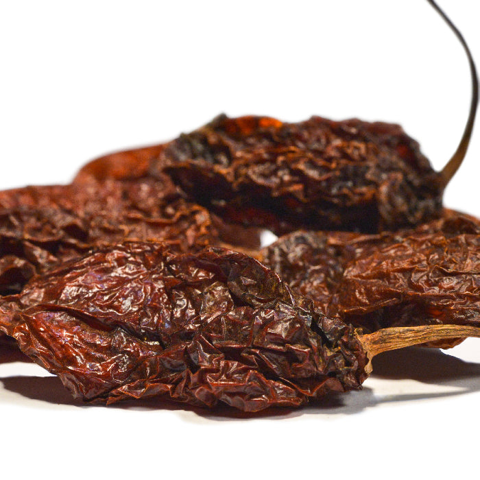A close-up of Whole Spice Inc. Ghost Chili Whole, one of the world's hottest peppers, shown next to dried chipotle peppers with wrinkled, dark reddish-brown skin against a white background.