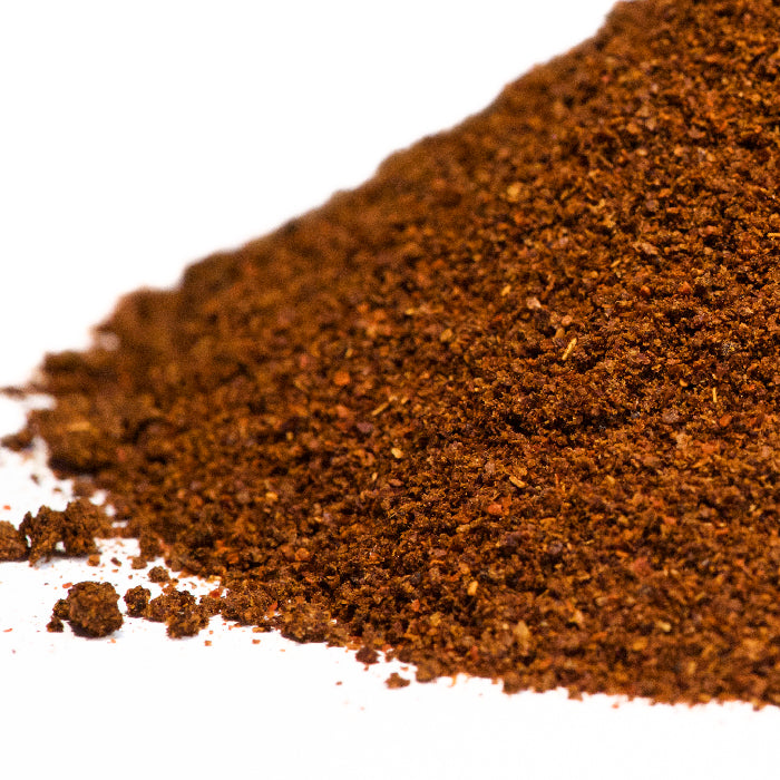 A close-up of Ghost Chili Powder from Whole Spice Inc., showcasing its finely ground, reddish-brown texture on a white surface; made from Bhut Jolokia, one of the world’s hottest peppers.