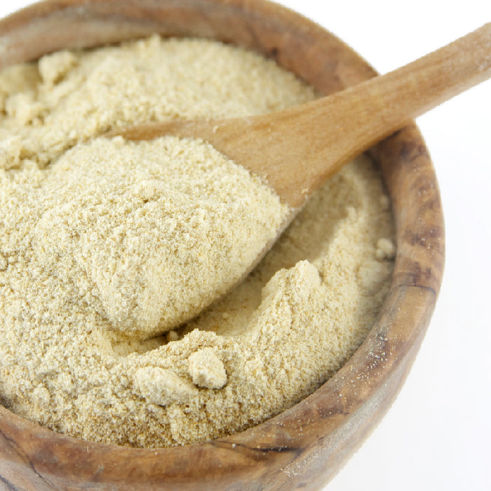 A wooden bowl filled with Whole Spice Inc. Ginger Salt, a light beige powder, with a wooden spoon scooping some inside. The background is white.