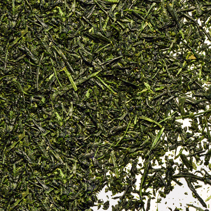 Close-up of loose Gyokuro Green Tea by Whole Spice Inc., with dried leaves scattered on a white surface, highlighting their deep green color and thin, irregular shapes.