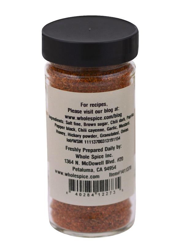 A clear spice jar with a black lid from Whole Spice Inc. displays a label for Honey BBQ Rub listing ingredients and company info. The reddish-brown sweet barbecue spice is visible, with a barcode at the bottom of the label.