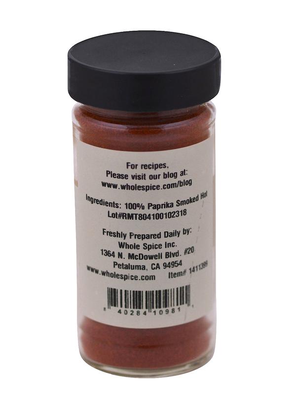 A clear spice jar with a black lid contains vibrant Hot Smoked Paprika by Whole Spice Inc., labeled as 100% smoked hot paprika, with company info and a website for recipe ideas.