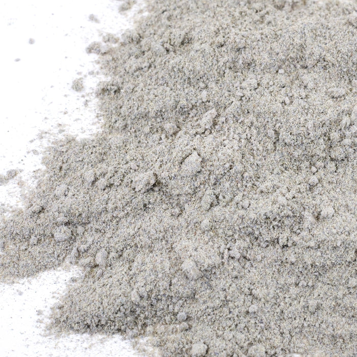 A close-up of Whole Spice Inc.'s Lavender Powder, a fine, light gray blend with a slightly uneven texture, spread on white and delicately flecked with dried lavender for an aromatic herbal appeal.