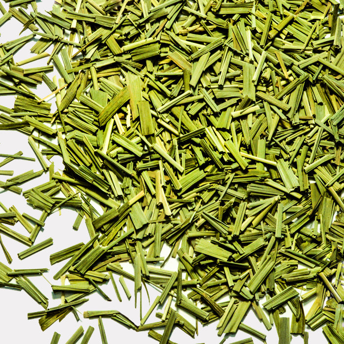 A close-up of Whole Spice Inc. Lemon Grass shows dried green pieces, cut into small, thin strips and scattered on a white surface—an essential Thai ingredient with a textured, layered look.