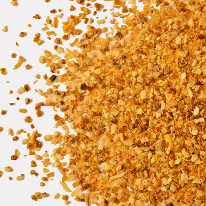 Close-up of Whole Spice Inc. Lemon Peel Granules scattered on a white background, highlighting their rough, golden-yellow texture—ideal for baking or pairing with dried garlic for an extra burst of zest.