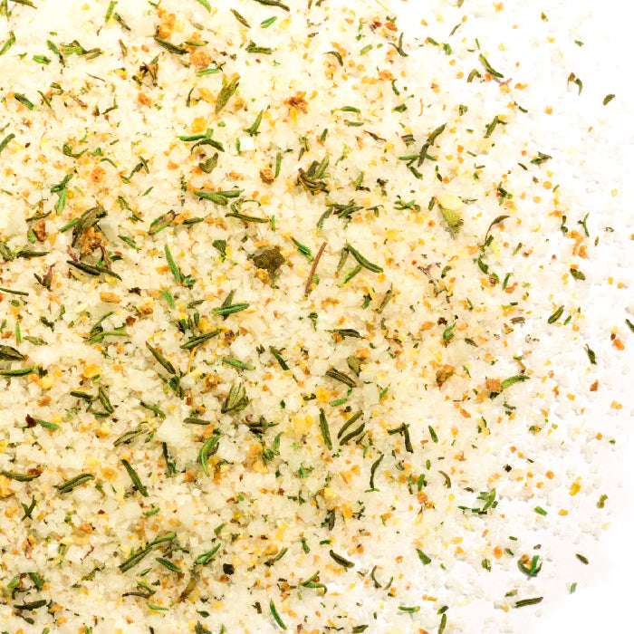 A close-up of Whole Spice Inc. Lemon Thyme Sea Salt, featuring coarse Fleur de Sel mixed with dried herbs and spices. Green, yellow, and brown flecks are scattered across a white surface.