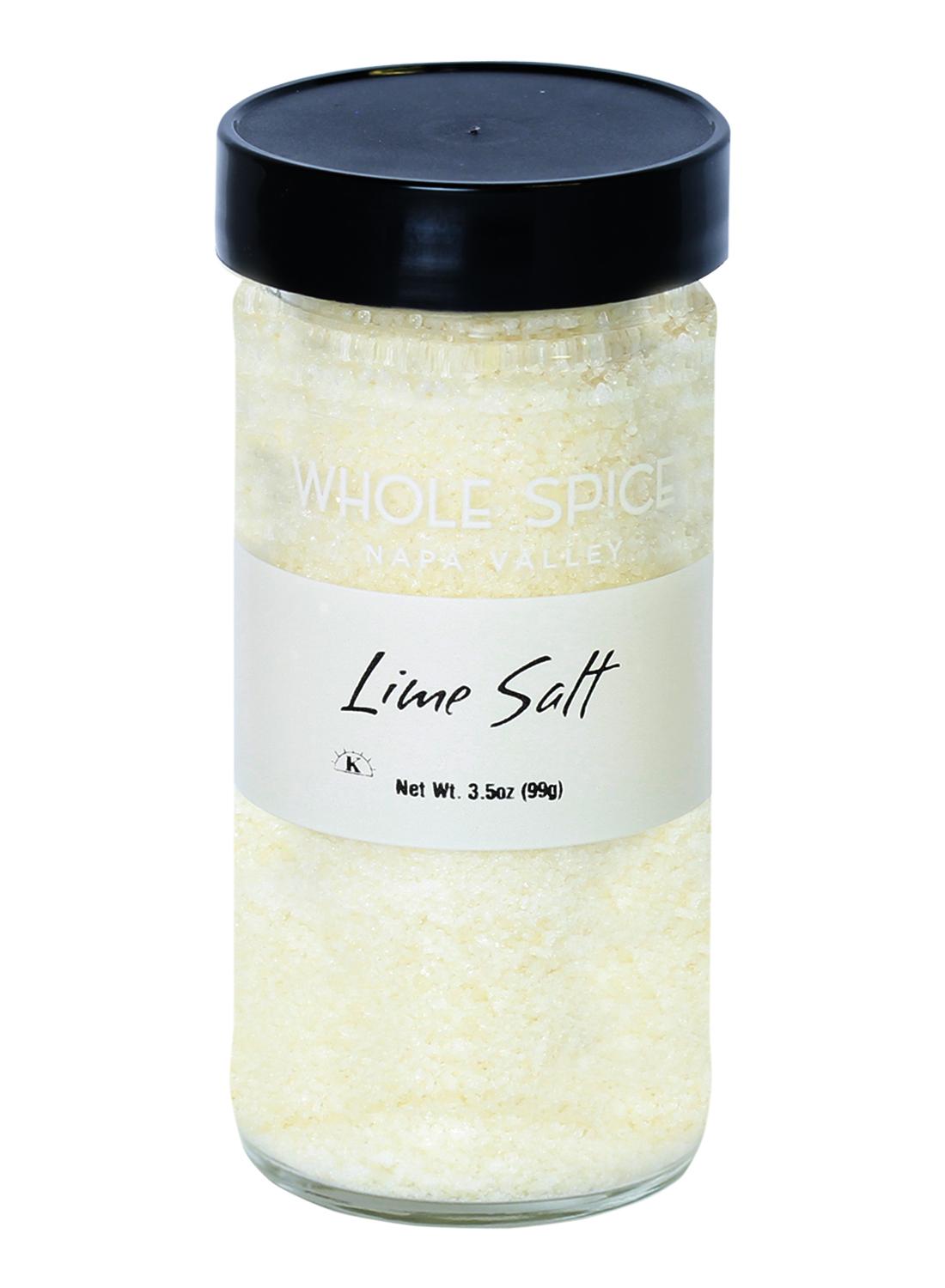 A clear glass jar with a black lid labeled “Whole Spice Inc. Lime Salt,” containing coarse, pale yellow sea salt with bright citrus flavor. Net weight: 3.5oz (99g).