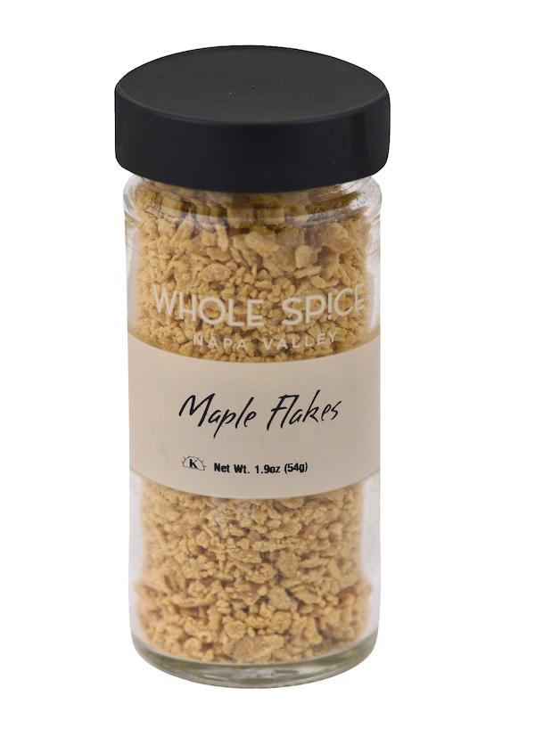 A clear glass jar with a black lid contains crunchy Maple Flakes from Whole Spice Inc., made from pure maple syrup. The label states "Maple Flakes" and notes a net weight of 1.9 oz (54g).