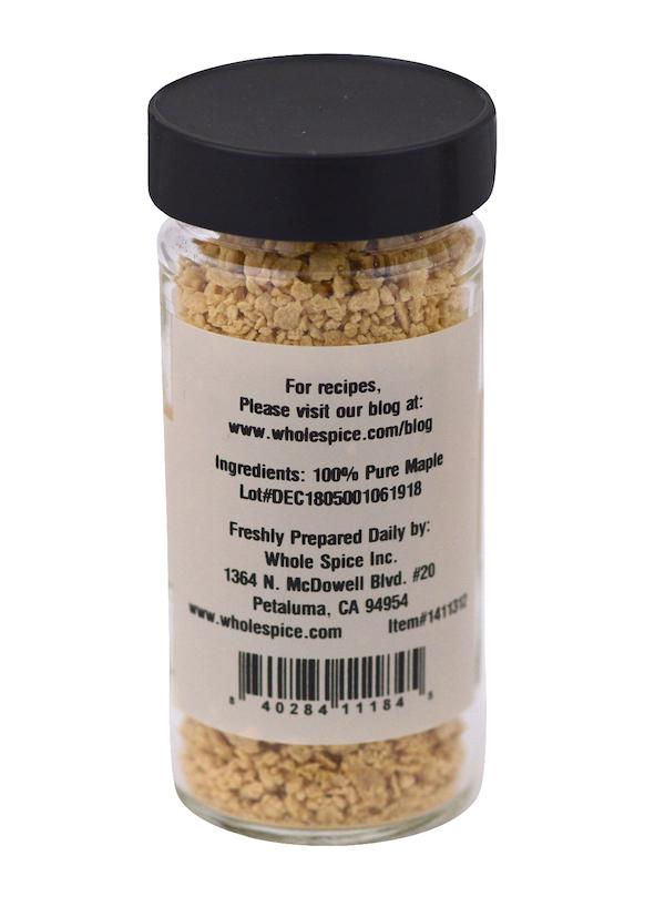 A clear glass jar with a black lid holds light brown crunchy Maple Flakes by Whole Spice Inc. The label lists 100% pure maple as the ingredient, displays company info, a recipe website, and features a barcode at the bottom.