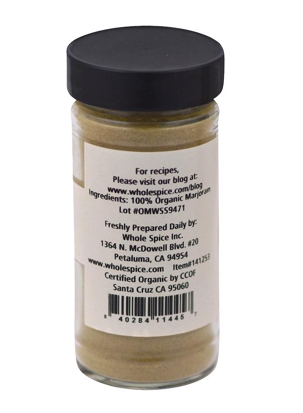 A glass jar with a black lid contains Marjoram Powder Organic from Whole Spice Inc. The label lists 100% organic herbs as the ingredient and includes Whole Spice’s contact info and certification details.