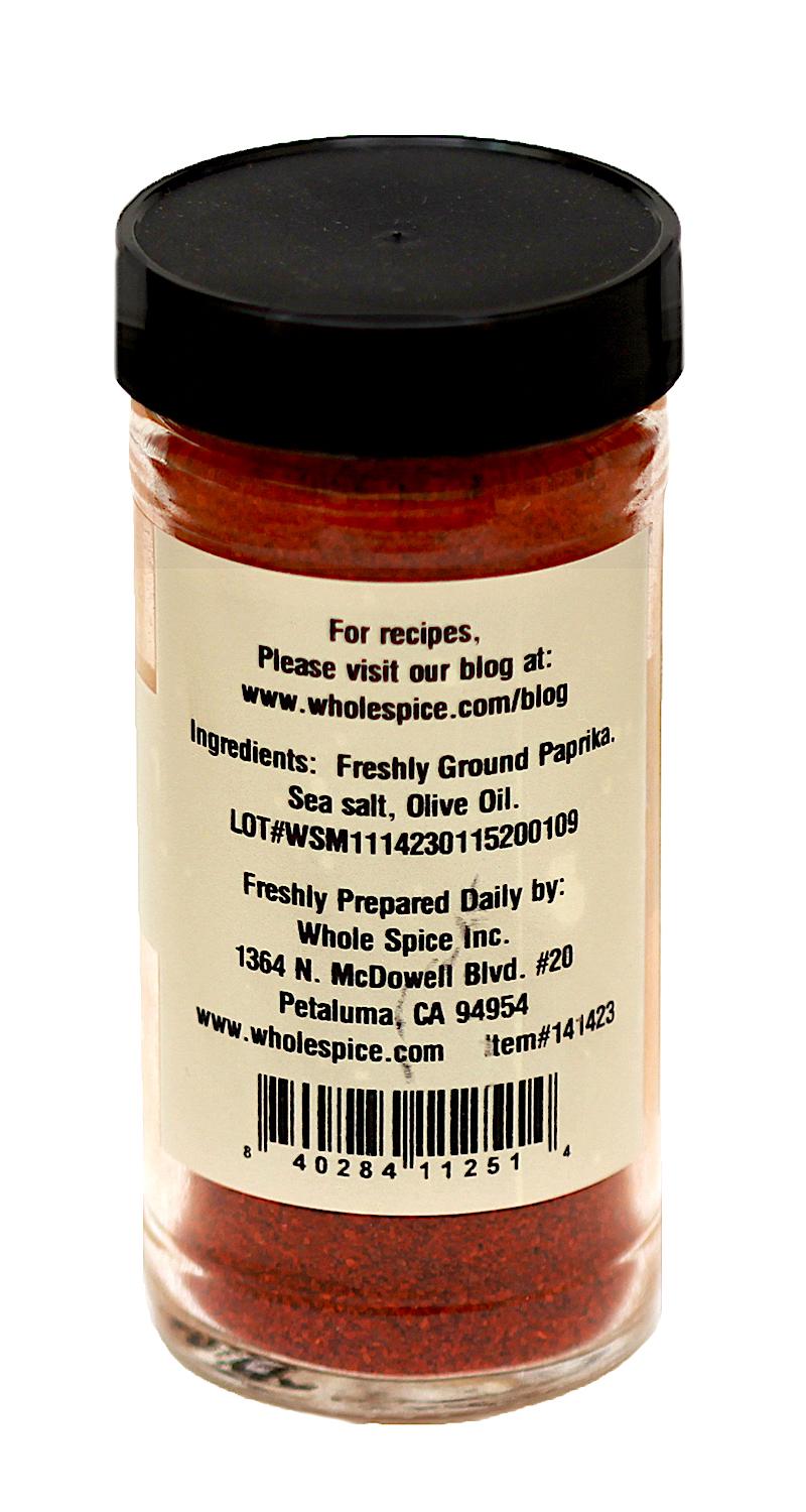 A clear spice jar with a black lid holds Whole Spice Inc.'s Moroccan Paprika & Olive Oil. The label lists freshly ground paprika, sea salt, and olive oil as ingredients, plus company and location details.