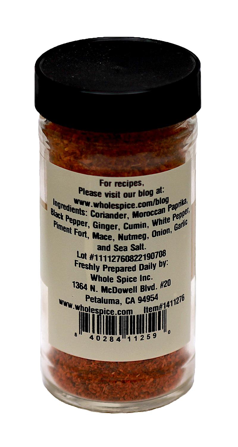 A clear spice jar with a black lid labeled "Moroccan Lamb Melange" by Whole Spice Inc. lists coriander, paprika, ginger, and sea salt. The label features website, address, barcode, prep directions—offering authentic Moroccan flavor.