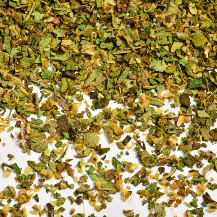 Close-up of Whole Spice Inc. Mexican Oregano leaves—coarse and flaky with green and brown hues—scattered on a white surface, highlighting this essential spice’s perfect seasoning for beans in authentic Mexican dishes.