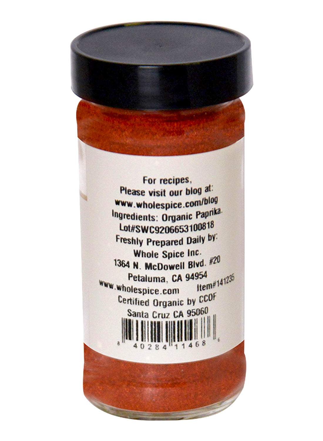 A clear spice jar with a black lid contains vibrant Paprika Organic by Whole Spice Inc. The label lists organic paprika as the main ingredient, along with certification, company address, a blog link, and a barcode.