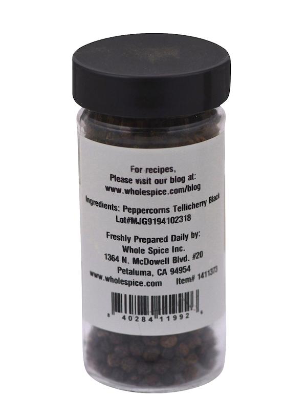 A clear jar labeled on the back contains Whole Spice Inc. Peppercorns Black Tellicherry with whole black peppercorns and company contact info from Petaluma, CA. Black lid included.