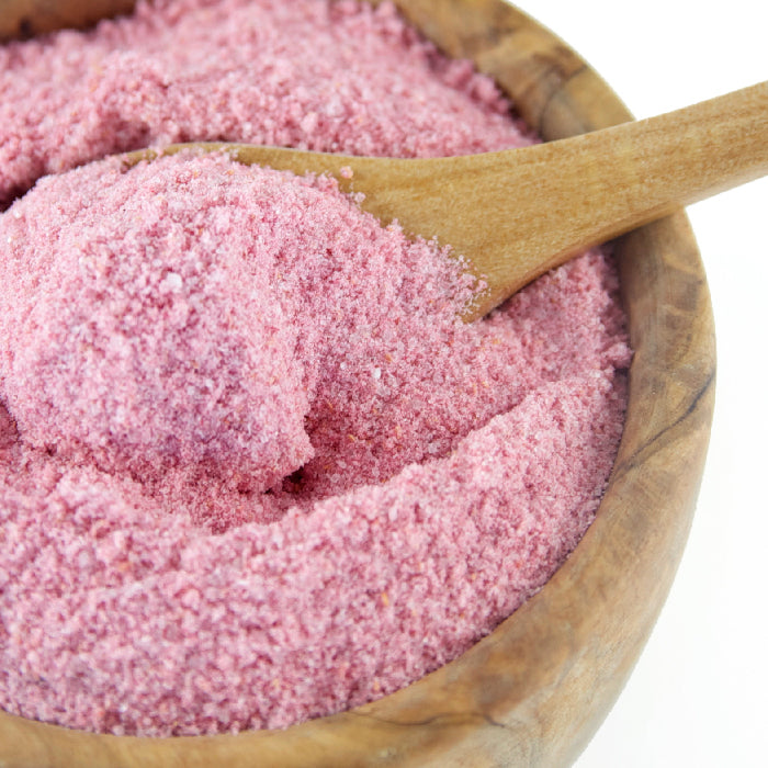 A wooden bowl holds Raspberry & White Chocolate Sugar by Whole Spice Inc., its fine pink, coarse grains scooped by a wooden spoon—ideal for adding gourmet flavor to white chocolate baking creations.