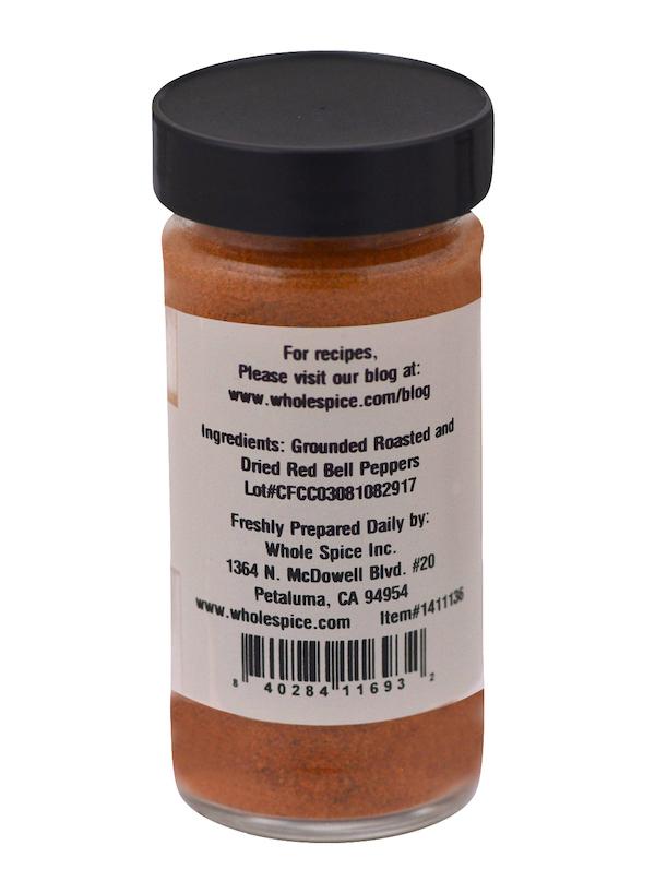 A clear spice jar with a black lid contains Roasted Red Bell Pepper Powder. The label from Whole Spice Inc. lists grounded roasted and dried red bell peppers as ingredients, along with company and manufacturing information.
