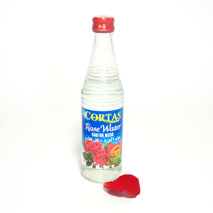 A clear 10 oz Rose Water Bottle from Whole Spice Inc. with a red cap and blue label stands upright, accompanied by a single red rose petal on a white background.
