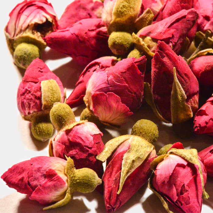 Close-up of Whole Spice Inc. Rosebuds with green sepals, scattered on a light surface. The tightly closed petals highlight their vibrant color and texture—ideal for adding fragrance or enhancing baking recipes.