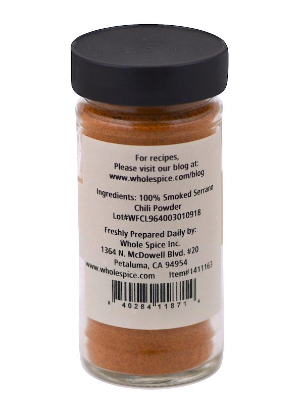 A clear jar with a sleek black cap contains vibrant orange-brown Smoked Serrano Chili Powder by Whole Spice Inc., labeled on the back with ingredients: 100% Smoked Serrano Chili Powder and contact details.