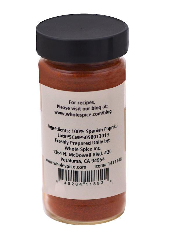 A clear spice jar with a black lid holds vibrant Spanish Paprika by Whole Spice Inc. The front label reads 100% Spanish Paprika, while ingredient, company, and contact information are printed on the back.