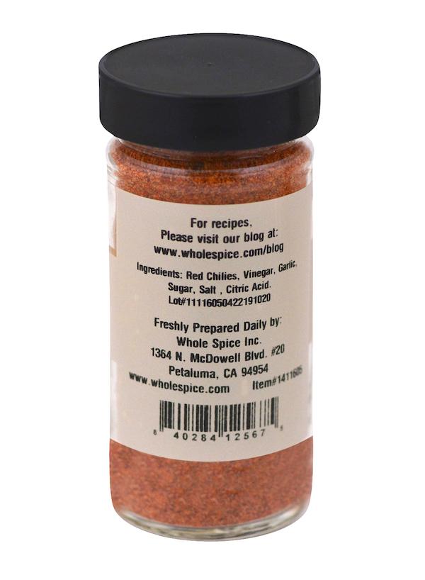 A clear spice jar with a black lid displays a label for Sriracha Hot Chili Seasoning by Whole Spice Inc., listing red chilies, vinegar, garlic, sugar, salt, and citric acid among the ingredients and providing contact info for Petaluma, CA.