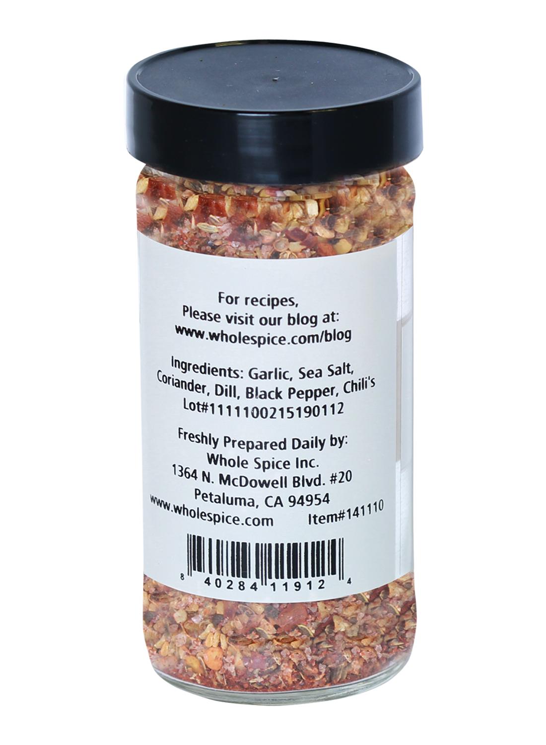 A clear spice jar with a black lid contains Whole Spice Inc. Steak Seasoning, a coarse blend of garlic, sea salt, coriander, dill, black pepper, and chili. The label shows preparation details and company information below the ingredients list.