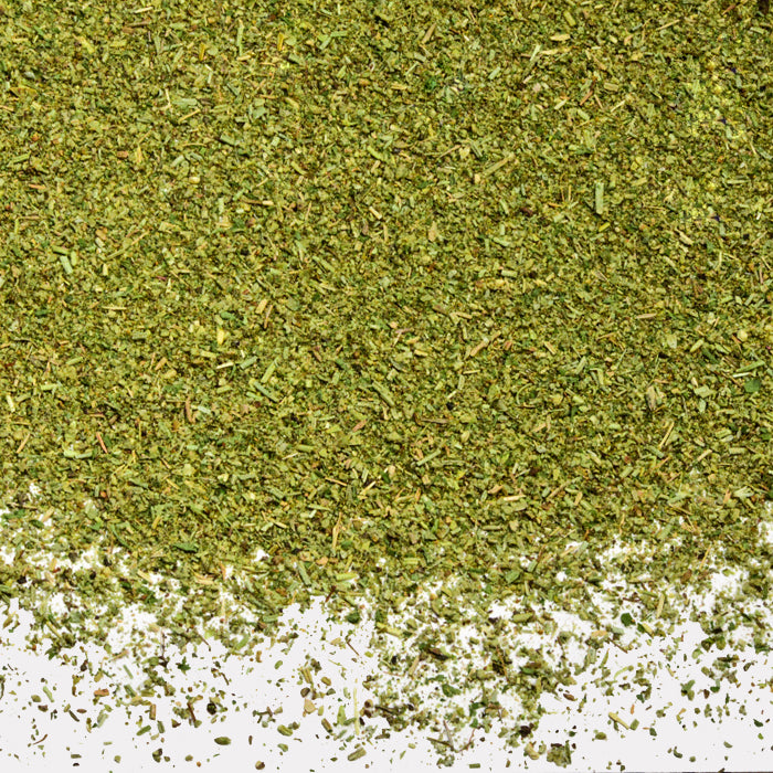 A close-up of Whole Spice Inc. Turkey Stuffing Mix, a blend of dried green herbs with visible fine and coarse pieces, scattered on a white surface and perfect for seasoning turkey dishes.