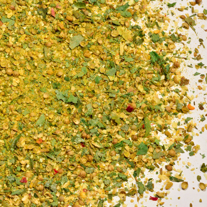 A close-up of Vadouvan Crushed by Whole Spice Inc.—a vibrant blend featuring green herbs, yellow powder, seeds, and dried vegetable pieces—showcasing the colorful ingredients found in this aromatic French-inspired spice mix.