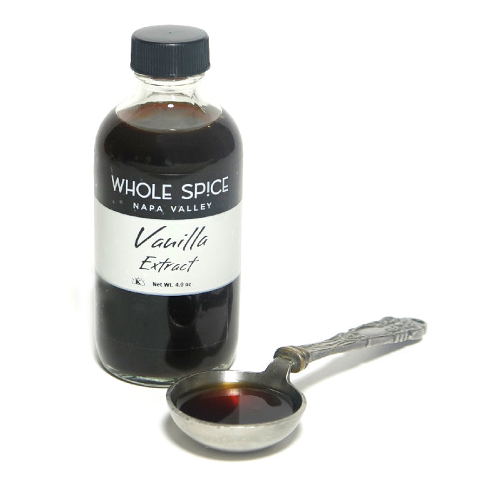 A glass bottle of Whole Spice Inc. Vanilla Extract sits beside a metal spoon of the dark vanilla extract, made from premium vanilla beans and ethyl alcohol, against a white background.
