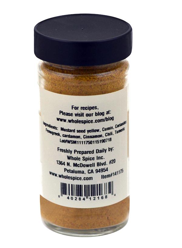 A clear jar with a black lid from Whole Spice Inc., labeled "Yellow Indian Curry Powder," contains a yellowish-brown blend of mustard seed, cumin, coriander, turmeric, plus company and website details.