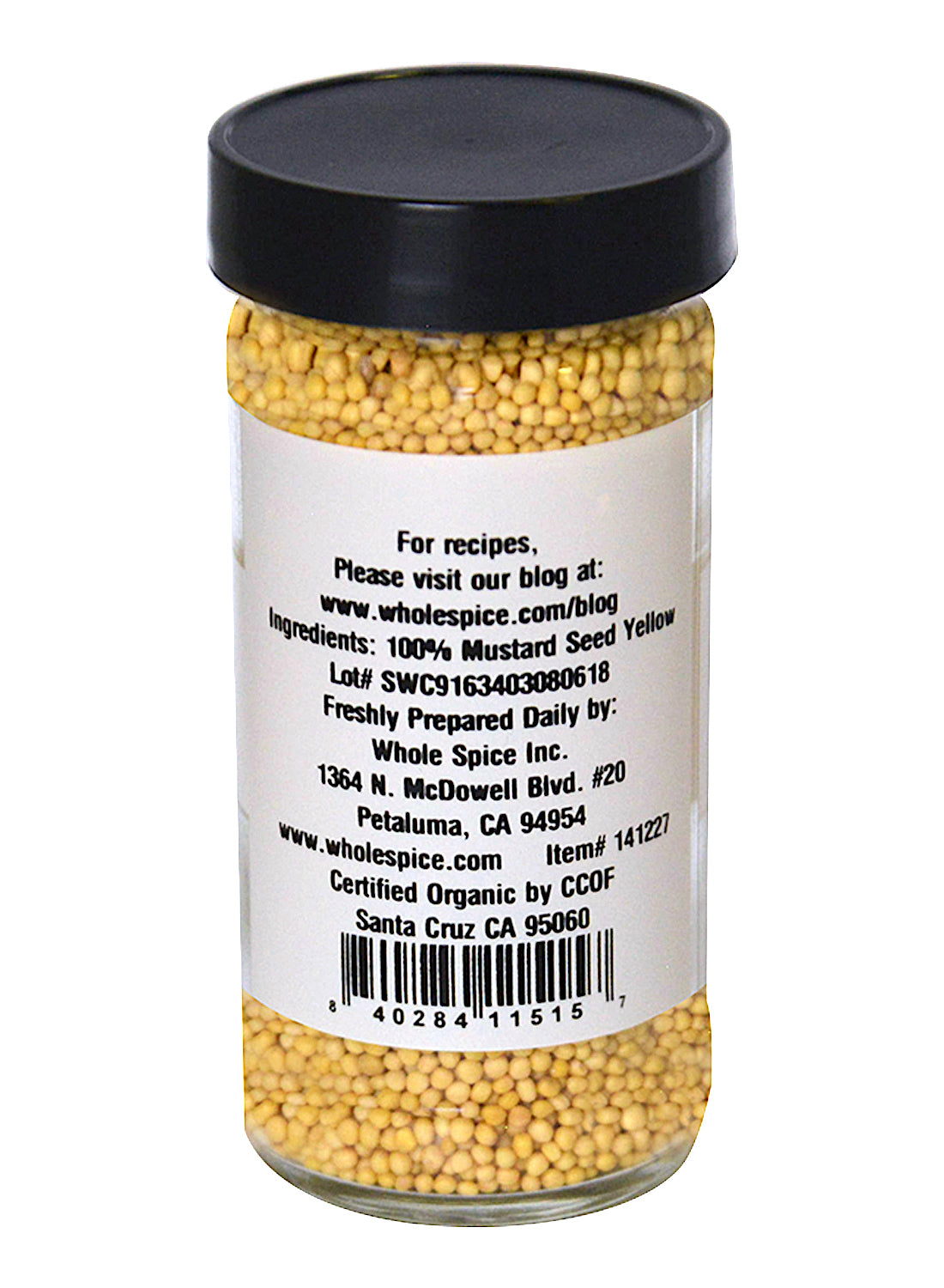 A clear glass jar with a black lid contains Whole Spice Inc. Yellow Mustard Seed Organic. The back label displays ingredients, company info, and a barcode.